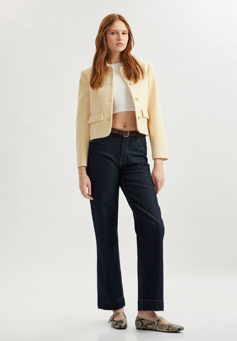 Cremfarbener Blazer mit goldenen Knöpfen über einem weißen, knappen Top, kombiniert mit dunkelblauen, geraden Jeans und Schlangenleder-Ballerinas.