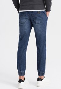 Joggers de mezclilla en azul oscuro con puños elásticos y un corte relajado, con bolsillos traseros y una pequeña etiqueta de marca en la cintura.