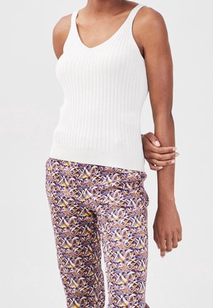 Femme portant un haut blanc sans manches en tissu côtelé et un pantalon aux motifs géométriques colorés, debout devant un fond uni.