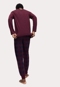 Personne portant une chemise à manches longues bordeaux et un pantalon de pyjama à carreaux rouge foncé et noir, vue de dos en train de tirer sur sa chemise.