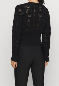 Svart stickad tröja med en vågmönster och öppna maskor, samt ribbade muddarna och nederkanten, tillsammans med åtsittande svarta byxor.