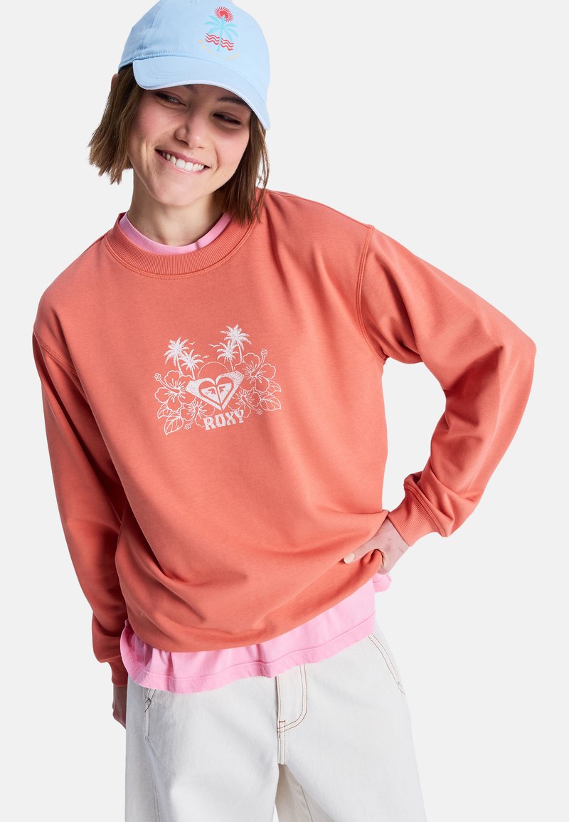 Korallenfarbener Sweatshirt mit weißem Blumenmotiv und "ROXY"-Logo, darüber ein pinkes Shirt getragen. Modell trägt eine hellblaue Kappe. Weicher Stoff, lockere Passform.