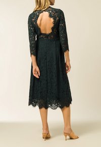 Donkergroene kanten jurk met driekwart mouwen, open rugontwerp en geschulpte zoom. Heeft ingewikkelde bloemenpatronen en knoopsluiting.