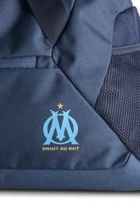 Sac de sport bleu marine avec des accents en maille, arborant un logo bleu clair et le texte doré "DROIT AU BUT" au-dessus d'une étoile. Matériau lisse et durable.