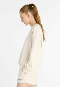 Sudadera color crema con hombros caídos y puños acanalados. Presenta un pequeño logo en el pecho y se lleva con pantalones cortos a juego.