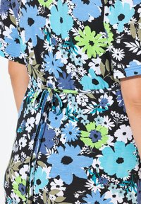 Kordovaný materiál šatů s velkými květinovými vzory v modré, zelené, bílé a černé barvě, svázaný tenkým páskem v pase, odhalující část paže osoby.