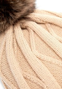 Cappello beige lavorato a maglia con motivo a treccia e un grande pon pon in pelliccia scura sulla sommità. Texture morbida e forma arrotondata.