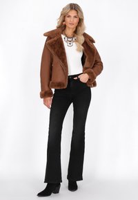 Giacca in finta pelliccia marrone con ampio colletto e polsini, abbinata a una maglia bianca e jeans flare neri. Ambientazione su sfondo bianco semplice.