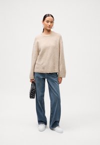 Béžový pletený sveter s okrúhlym výstrihom, kombinovaný s voľnými modrými džínsy a bielymi teniskami. Drží čiernu tašku so striebornými cvokmi.