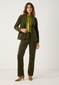 Olivgrünes Cordblazer und passende Hose, kombiniert mit einem hellgrünen Satinhemd. Braune spitze High Heels vervollständigen das Outfit.