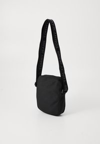 Svart skulderveske med rektangulær form, laget av teksturert stoff, med en tykk stropp med "TOMMY JEANS"-logo. Glidelås luke.