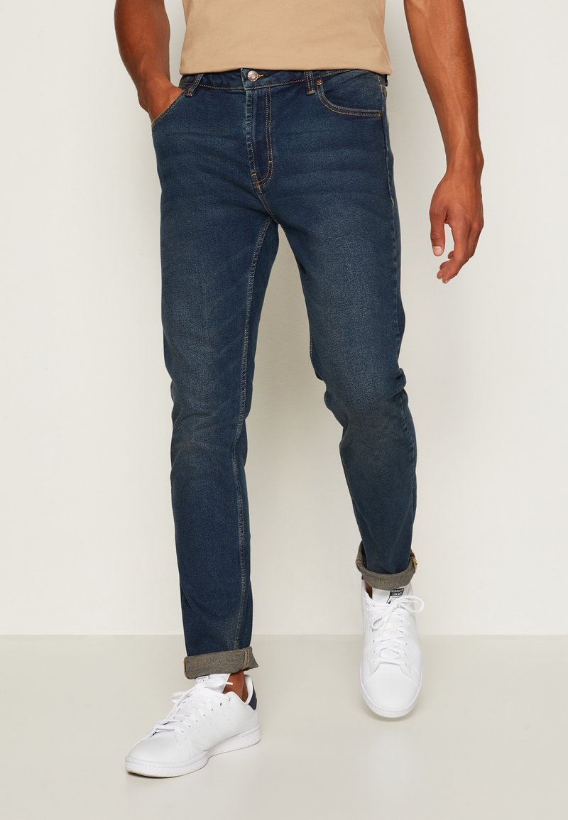 Mörkblå jeans med slim fit, med standardfickor, knappknäppning och uppvikta fållar. Bärs med vita sneakers.