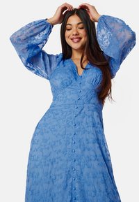 Blå broderad klänning med långa puffärmar, knappstängning framtill, blommönster och textil med struktur. Elegant silhuett med figursydd överdel.