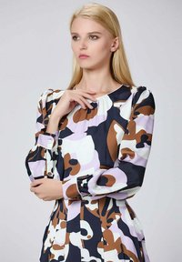 Femme blonde aux cheveux raides portant une robe à manches longues avec des motifs abstraits marron, noir, blanc et lavande, posant avec une main près de son visage.