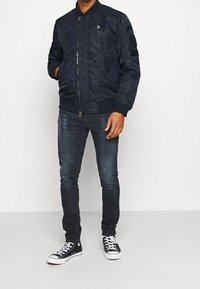 Marinblå bomberjacka med dragkedja fram, ribbade ärmar och två sidofickor; har en quiltad design och märken på ärmarna.