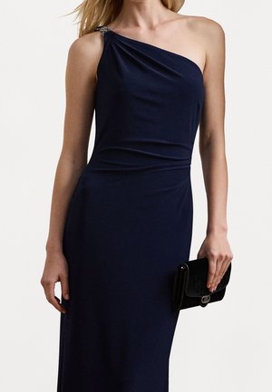 Kvinde iført en ærmeløs marineblå one-shoulder kjole, der holder en lille sort clutch taske ved siden af sig.