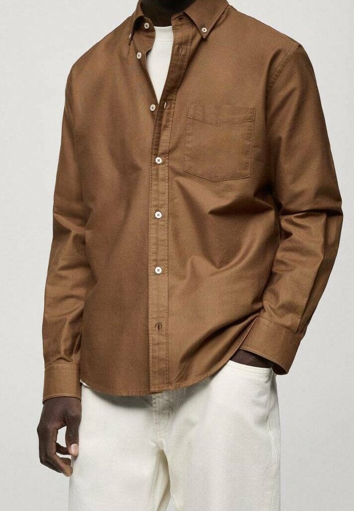Homme portant une chemise marron à boutons par-dessus un t-shirt blanc, avec un pantalon beige clair, main dans la poche, visage non visible.
