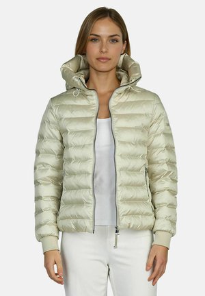 Frau trägt eine hellbeige gesteppte Steppjacke mit Kapuze über einem weißen Oberteil und weißen Hosen, steht vor einem schlichten Hintergrund.