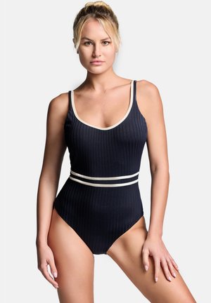Donna bionda che indossa un costume da bagno intero nero a coste con bordi bianchi e doppie strisce bianche in vita, in piedi con le braccia rilassate.