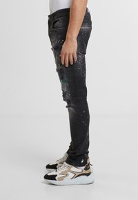 Cienkie czarne dżinsy z przetarciami, efektem splamienia farbą i wyblakłą teksturą. Połączone z beżowymi i czarnymi trampkami.