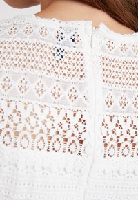 Biała koronkowa bluzka z misternymi wzorami wyciętymi, teksturowanymi warstwami oraz ukrytym zamkiem błyskawicznym z tyłu, prezentująca szczegółowe rzemiosło.
