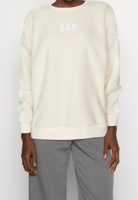 Polarowa bluza w kolorze kremowym, z okrągłym dekoltem, ściągaczami na mankietach i u dołu; na piersi znajduje się haftowane logo "GAP" w kolorze białym.