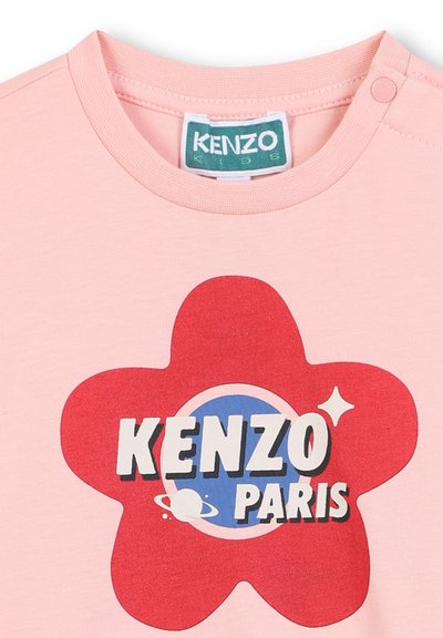 Różowa bawełniana koszulka z nadrukiem czerwonego kwiatu na niebieskim kole, z białym napisem "KENZO PARIS" oraz zapięciem na zatrzask przy ramieniu.