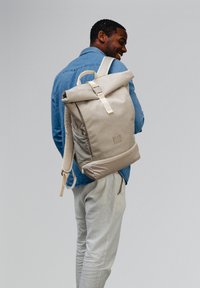 Beige roll-top backpack made of durable fabric, featuring padded straps, a front zip pocket, and a minimal logo accent.