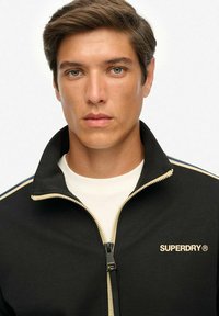 Chaqueta negra con cremallera y cuello alto, con detalles dorados y logo. La tela parece suave con franjas beige contrastantes en los brazos.