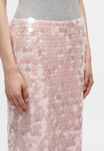 Gros plan d'une personne portant un haut beige côtelé et une jupe en filet rose transparent avec des sequins irisés, les mains reposant le long du corps.