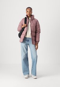 Rosa puffer-jakke med høy krage, båret over en kremfarget kabelstrikket genser. Lys blå vide jeans og en svart skulderveske. Hvite sneakers.