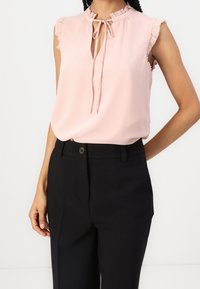Blouse sans manches rose pâle avec un col à volants et un détail en forme de goutte, associée à un pantalon noir sur mesure, mettant en valeur un tissu lisse et une coupe décontractée.