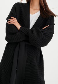 Musta ribattu neuletakki, jossa on pyöreä pääntie, pitkät hihat ja solmittava vyötärö. Materiaali näyttää pehmeältä ja teksturoidulta.