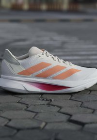 Chaussure de course blanche avec des bandes orange, un support de talon gris et un amorti rose, posée sur un sol en pierre texturé.