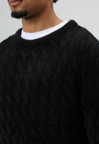 Zwarte kabelgebreide trui met een zachte, pluizige textuur en een brede ronde hals, gedragen over een wit overhemd. Close-up van de stofdetails.