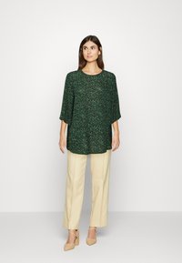 Blusa floral verde com um corte descontraído, mangas 3/4, decote redondo, combinada com calças largas creme e sapatos com salto bloco em tom nude.