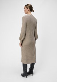 Lange beige gebreide jurk met een geribde textuur, ballonmouwen en subtiele naaddetails. Draagt zwarte puntige haklaarzen.