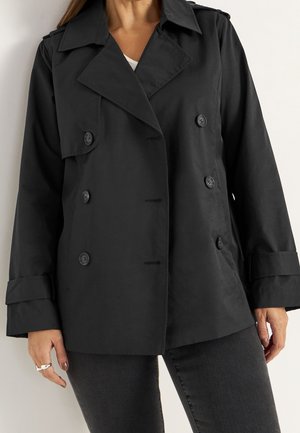 Femme portant une veste noire à double boutonnage avec de larges revers et des détails de boutons, associée à un pantalon foncé et une chemise blanche en dessous.