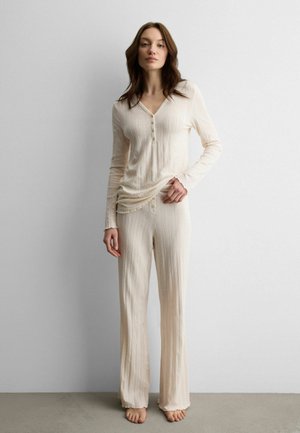 Frau steht barfuß und trägt ein cremefarbenes, geripptes Langarmshirt und passende weite Hosen vor einem einfachen hellen Hintergrund.