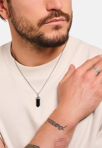 Silberne Halskette mit einem schwarzen Kristallanhänger, getragen von einer Person in einem beigen Hemd. Die Hand zeigt einen strukturierten Ring und sichtbare Tätowierungen.