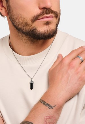 Silver necklace with a black crystal pendant, worn by a person in a beige shirt. Hand showcases a textured ring and visible tattoos.