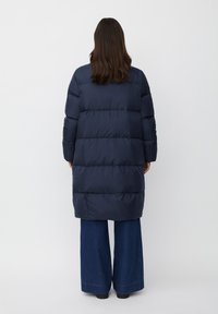 Cappotto imbottito blu navy con design lungo, trapuntatura orizzontale e colletto alto. Abbinato a pantaloni in denim a gamba larga.