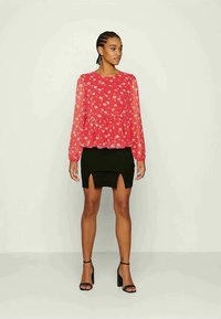 Blusa vermelha floral com mangas longas transparentes, combinada com uma saia preta justa com uma abertura frontal. Modelo usa sandálias pretas de salto.