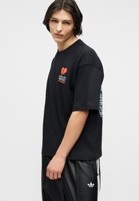 Νεαρός άνδρας που φοράει μαύρο γραφικό μπλουζάκι με κόκκινη σπασμένη καρδιά και μαύρο παντελόνι Adidas σε απλό λευκό φόντο.