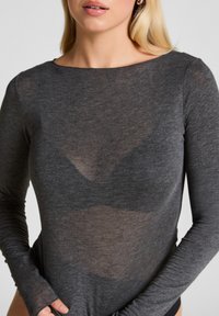 Vrouw in een doorzichtige, lange mouwen grijze top die een donkere bh eronder onthult, met handen die de onderrand van het shirt vasthouden.