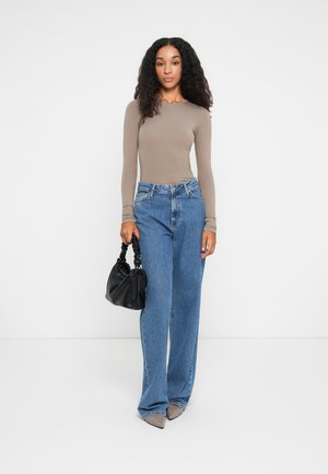 Mujer con cabello rizado, vestida con una blusa de manga larga beige, jeans de pierna ancha azules, zapatos gris puntiagudos, sosteniendo un bolso negro fruncido, de pie contra un fondo blanco.