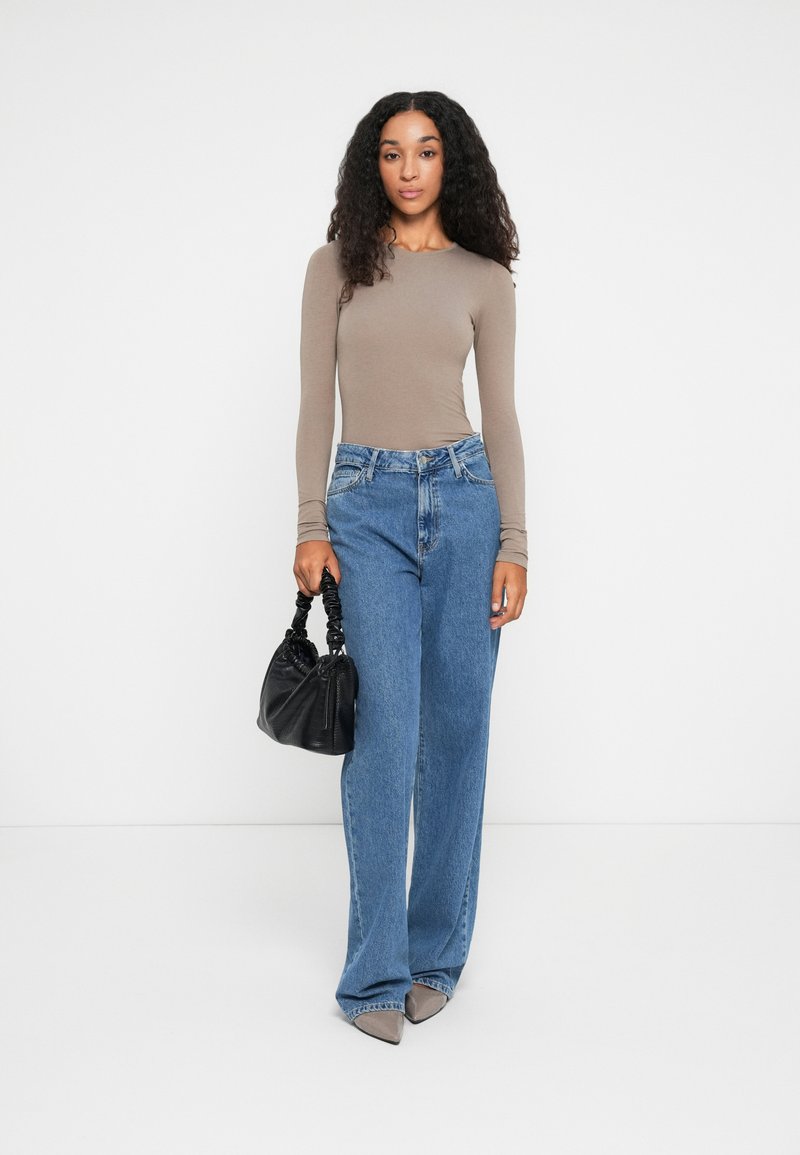 Kvinne med krøllete hår som har på seg en beige langermet topp, blå vide jeans, grå spisse sko, holder en svart rynket håndveske, stående mot en hvit bakgrunn.