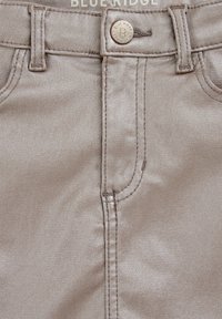 Beige színű, selyemfényű farmernadrág, elülső gombos záródással, fém kiegészítőkkel, övhurkokkal és varrási részletekkel. A márka logója a derékpánt felett látható.