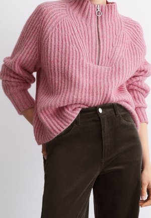 Woman wearing a ribbed pink quarter-zip sweater tucked into high-waisted dark brown corduroy pants with hands resting near pockets.