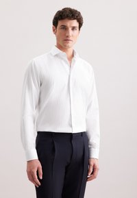Camicia bianca a maniche lunghe con righe verticali, colletto button-down e polsini, abbinata a pantaloni eleganti blu navy. Texture del tessuto liscia.